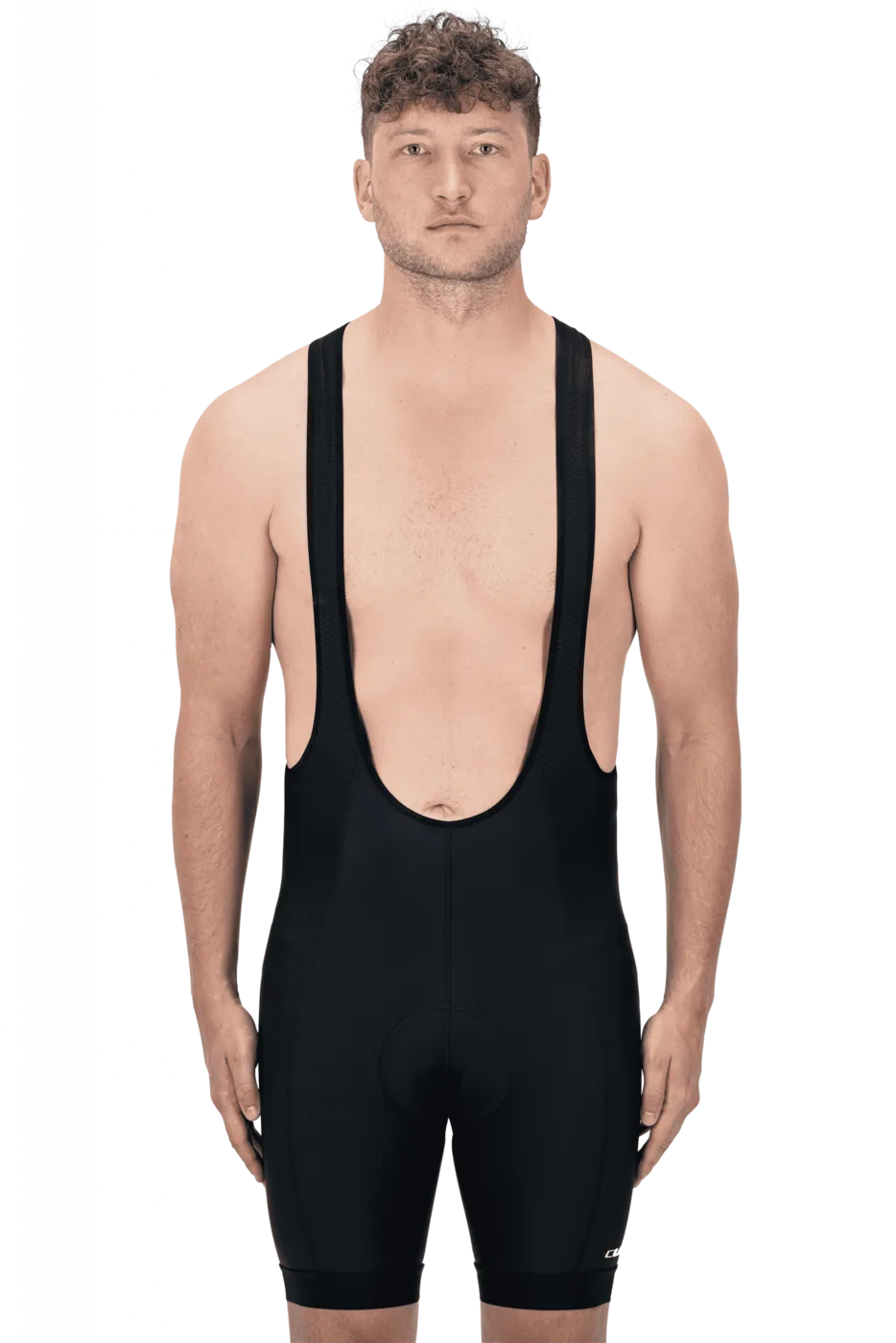 Ein Mann mit kurzen lockigen Haaren und ohne Hemd steht in einer CUBE ATX Trägerhose neben seinem Fahrrad, mit neutralem Gesichtsausdruck auf schlichtem Hintergrund, bereit für eine Fahrt um Lichtenfels.