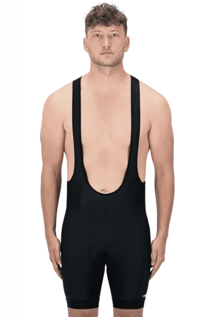 Ein Mann mit kurzen lockigen Haaren und ohne Hemd steht in einer CUBE ATX Trägerhose neben seinem Fahrrad, mit neutralem Gesichtsausdruck auf schlichtem Hintergrund, bereit für eine Fahrt um Lichtenfels.